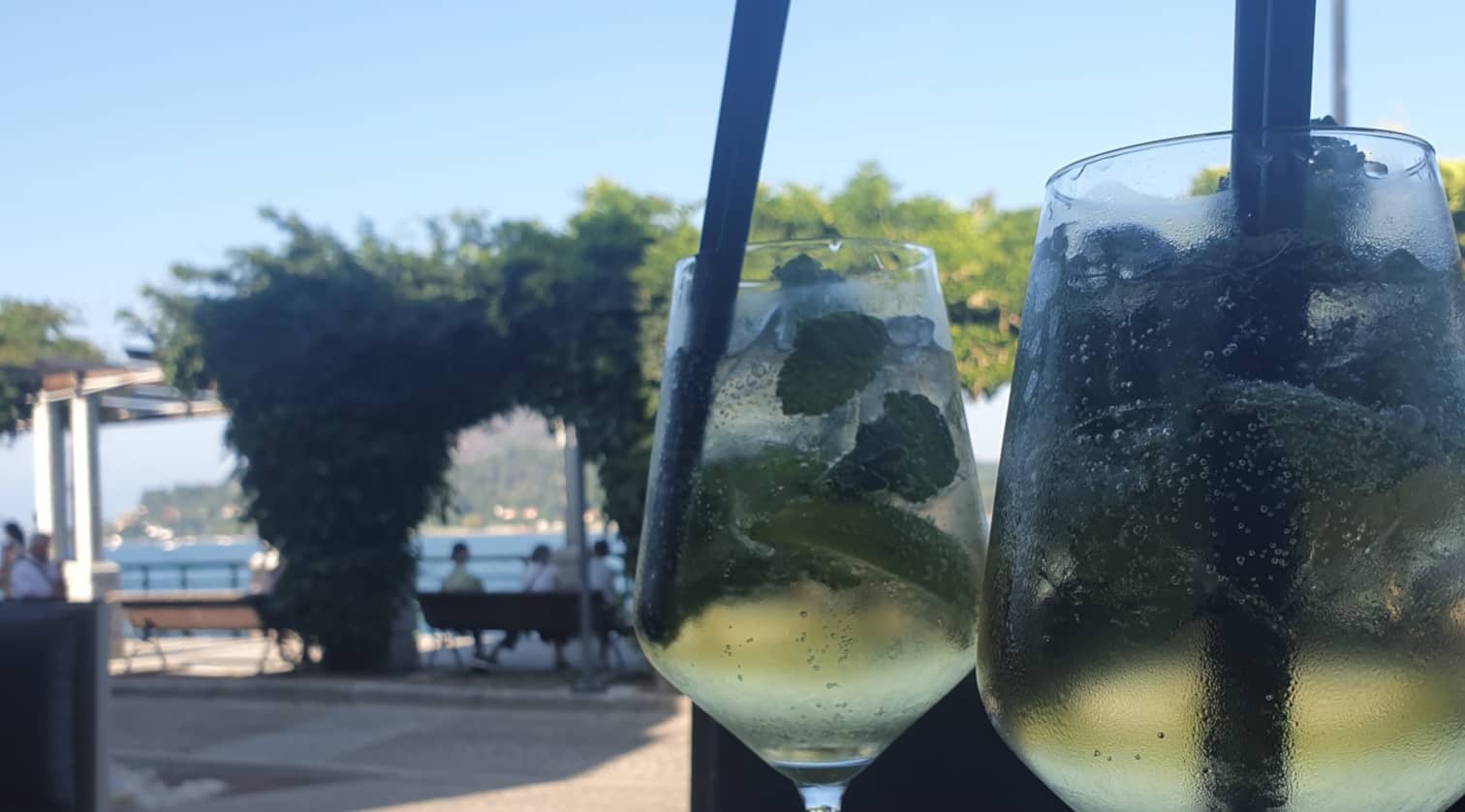 Gros plan à contre-jour : menthe, citron vert et glaçons derrière la buée des verres ; en arrière-plan flou, des bancs occupés le long de l'eau.