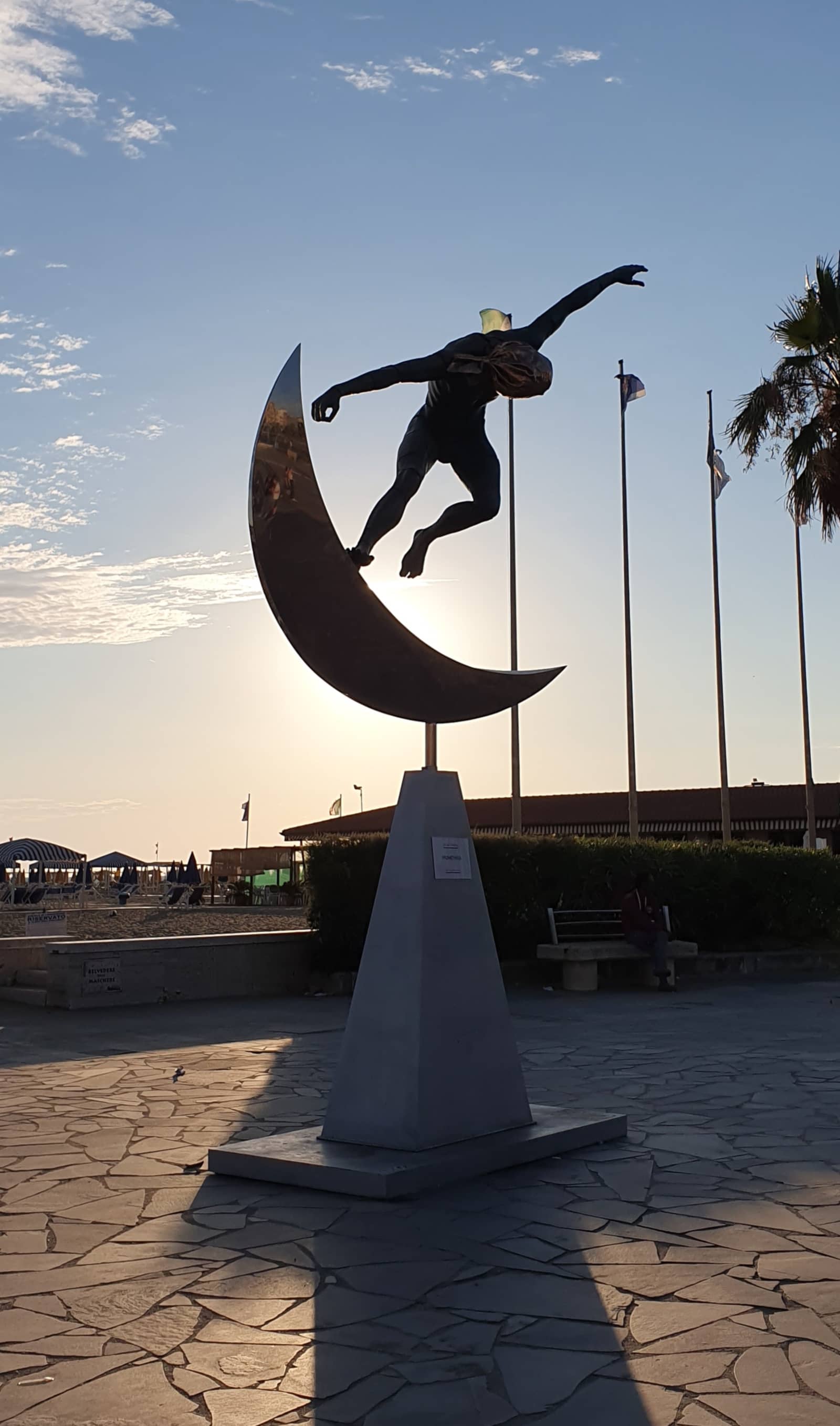 Bronze sculpture of a figure balancing on a crescent moon, backlit at sunset on the Viareggio seafront.
